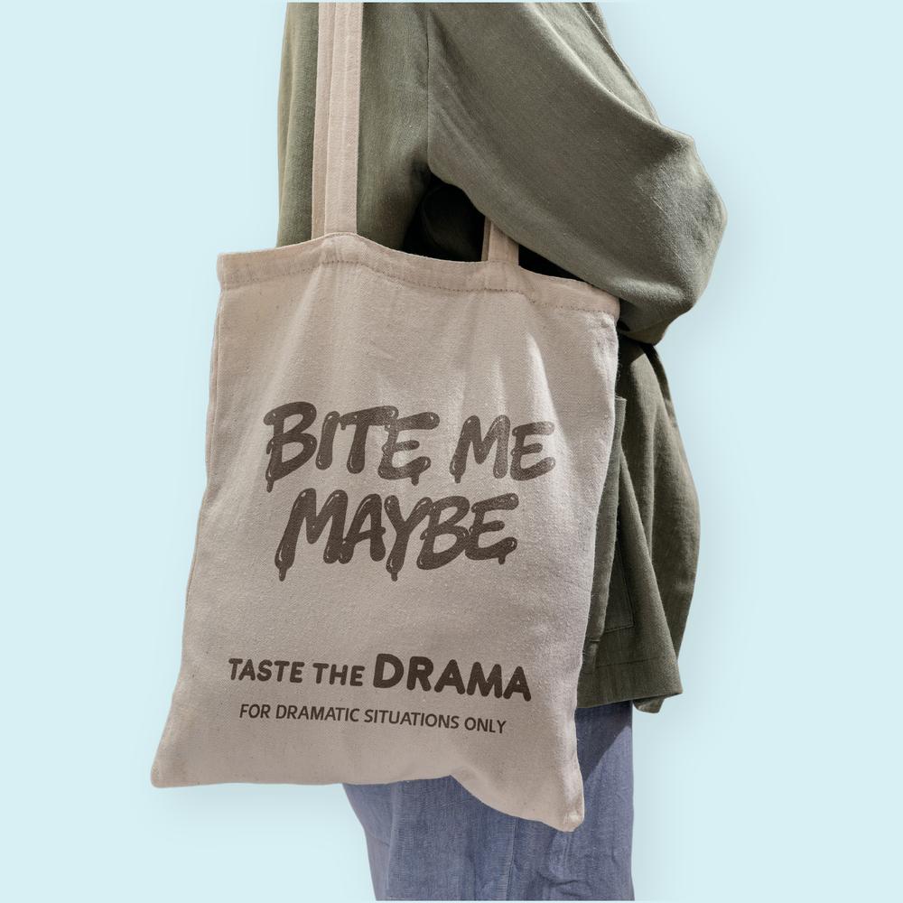 A person wearing an oversized grey tote bag printed with the large "Bite Me Maybe" logo and branding text.