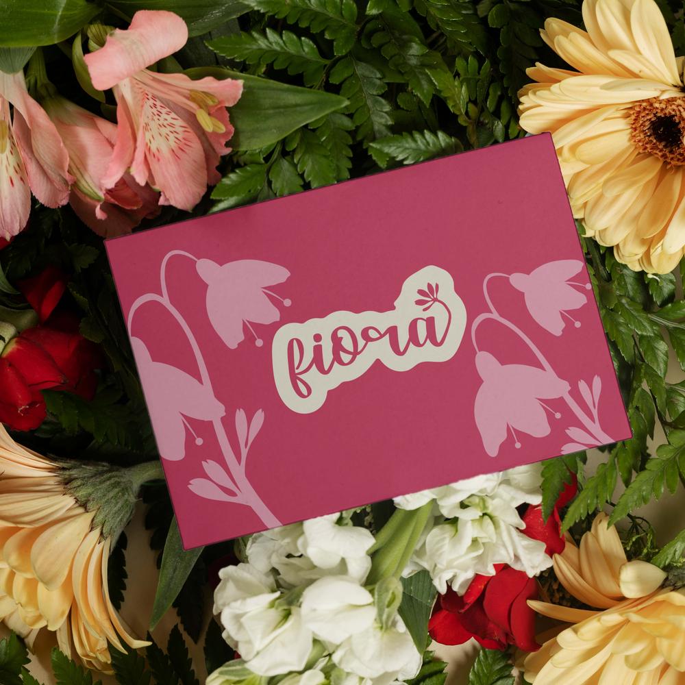 A burgundy Fiora branded gift envelope resting on a bed of yellow gerbera daisies and red roses.